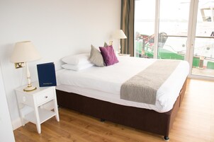 Habitación de hotel con cama, dos mesitas de noche, lámpara, un libro y vistas a barcos a través de grandes ventanales.