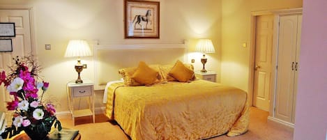 Un dormitorio con una cama grande, dos mesitas de noche con lámparas, una mesilla auxiliar, un jarrón de flores y un cuadro en la pared.