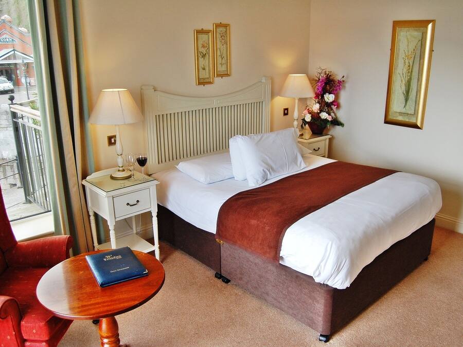 A hotel room with a bed, a red armchair, a small table with a book, a nightstand with a lamp, and a view of the street outside.