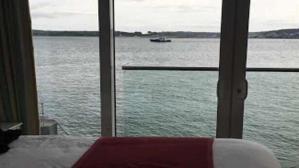 A room with a bed, a red blanket, and a view of a body of water with a boat. - WatersEdge Hotel (Cobh)