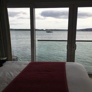 A room with a bed, a red blanket, and a view of a body of water with a boat. - WatersEdge Hotel (Cobh)