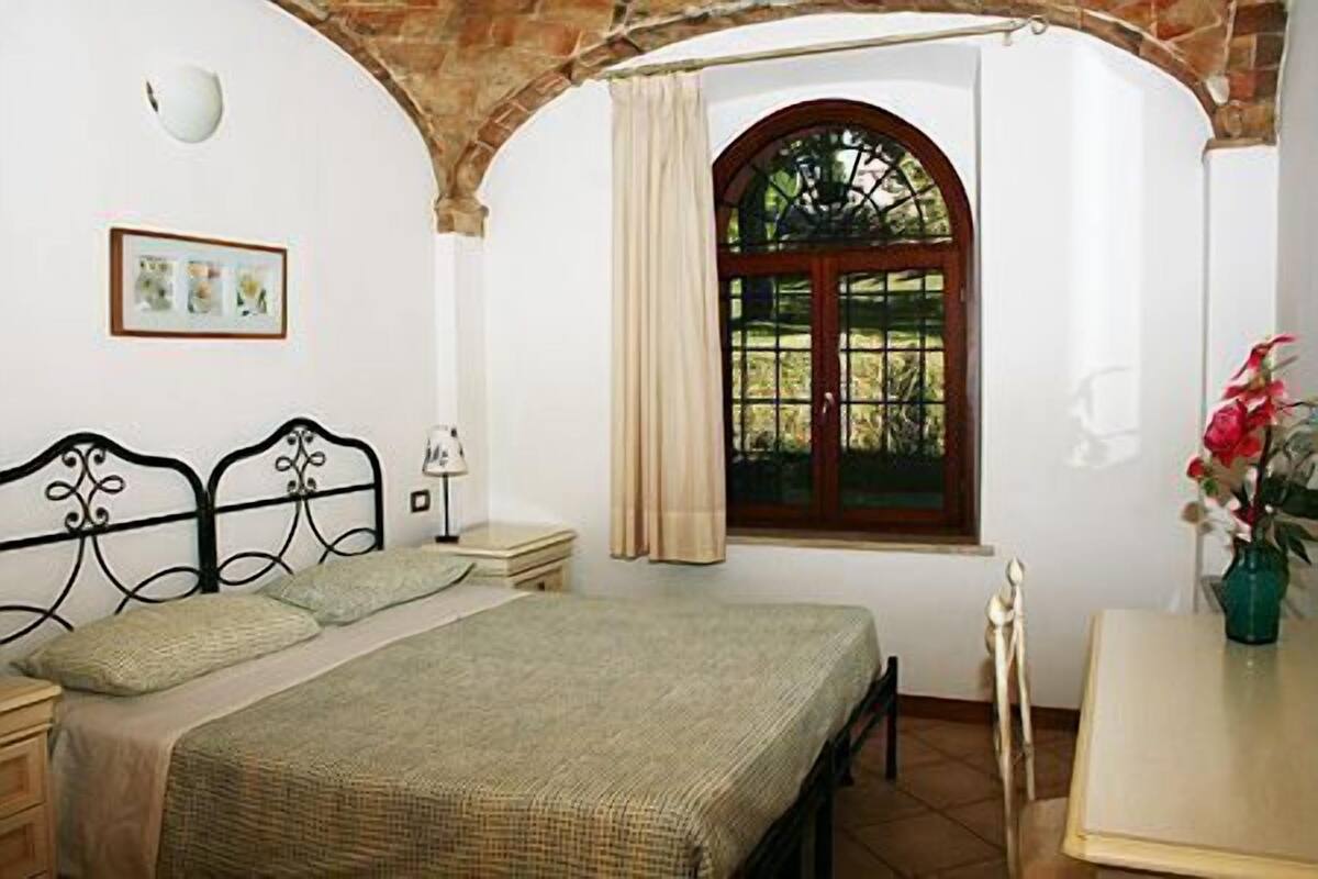 A bedroom with a bed, a nightstand, a lamp, a window with curtains, and a vase with flowers.