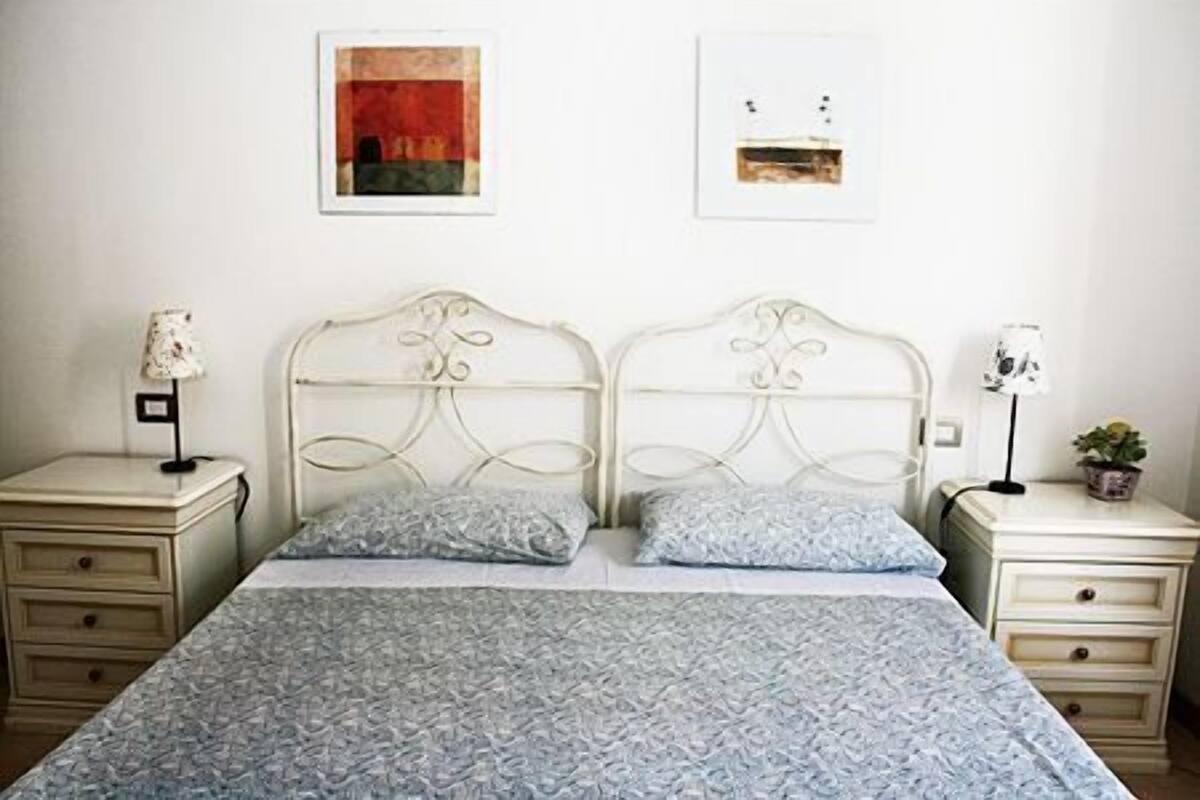 A double bed with a patterned bedspread, two bedside tables with lamps, and two framed pictures on the wall.