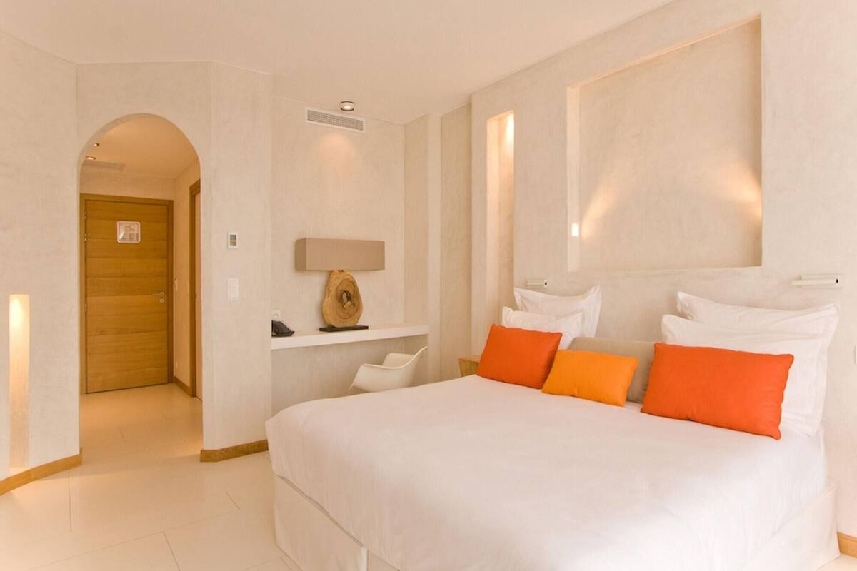 A hotel room with a large bed, a wooden door, a small desk with a lamp, and a wall-mounted shelf.