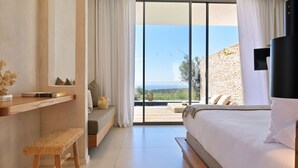 A bedroom with a large bed, a wooden bench, a desk, and a view of the outdoors through large windows. - Hotel Cala di Greco (Bonifacio)