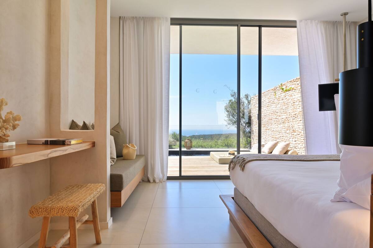 A bedroom with a large bed, a wooden bench, a desk, and a view of the outdoors through large windows.