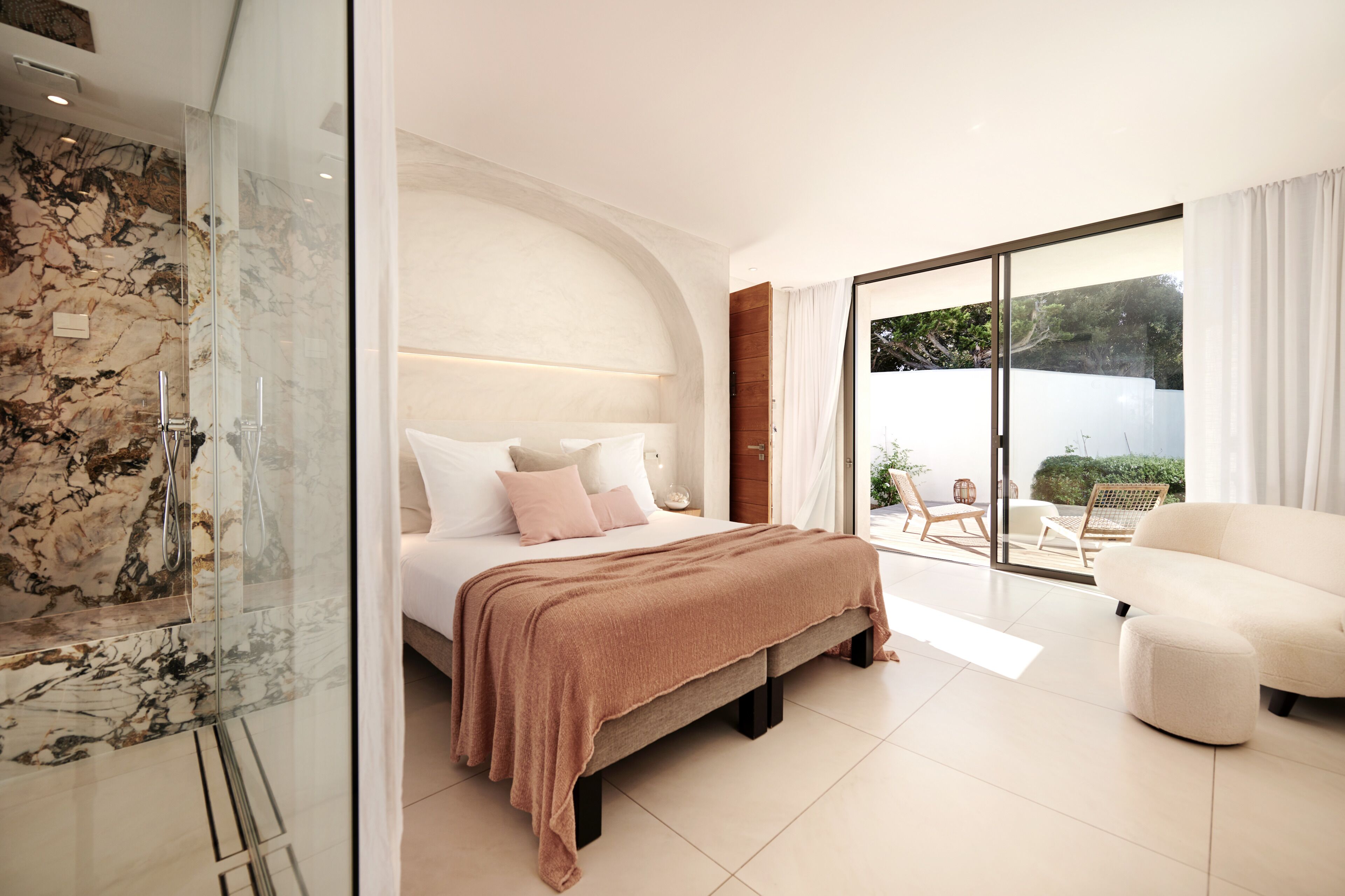 A modern bedroom with a large bed, a glass shower enclosure, and a balcony with outdoor seating.