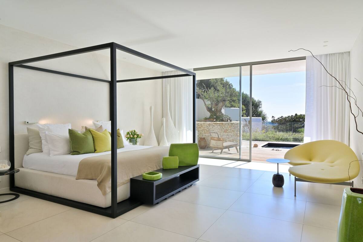 A modern bedroom with a four-poster bed, a green accent chair, and a view of the outdoors.