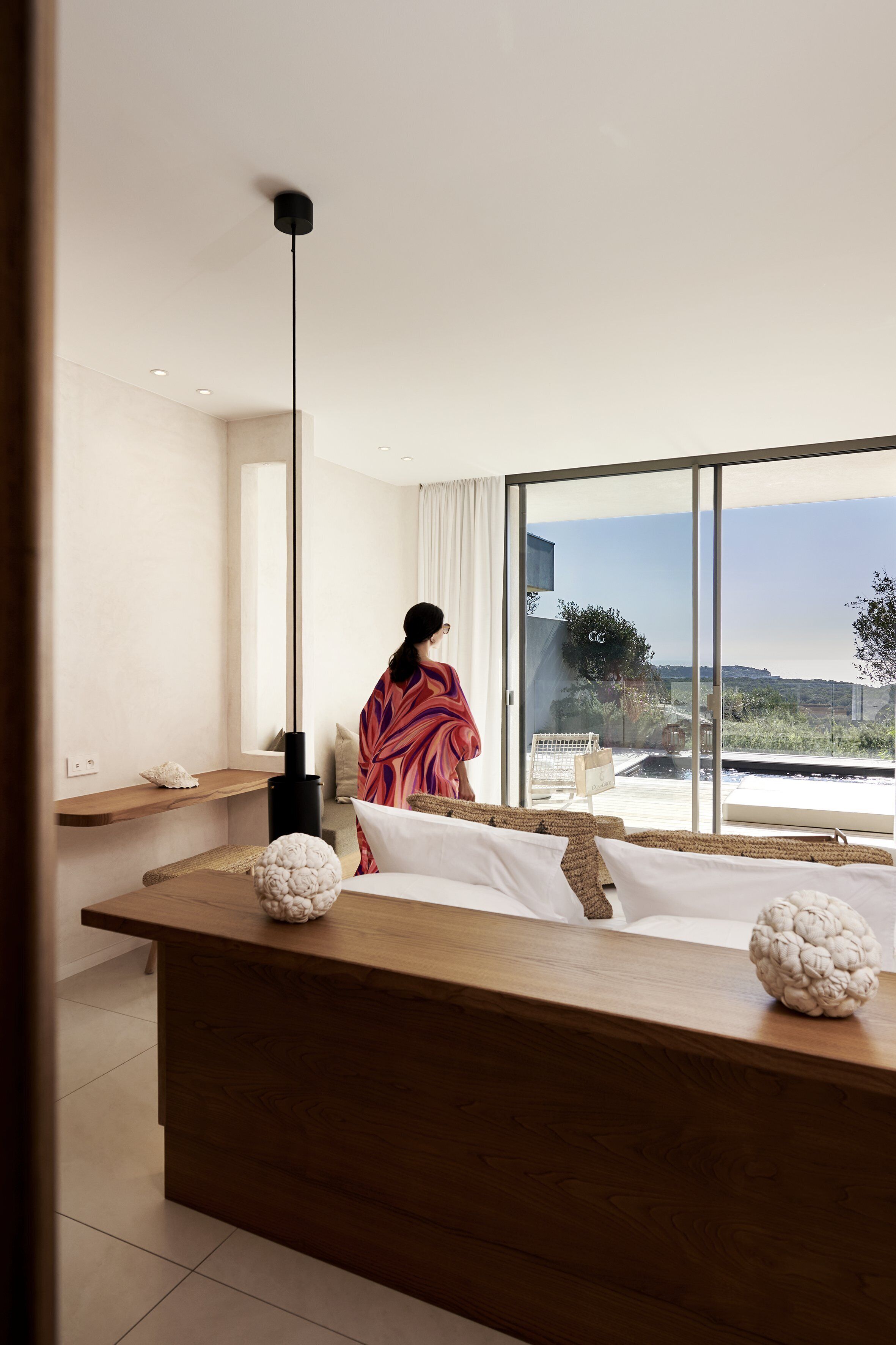 A modern bedroom with a large window, a wooden bedside table, and a person in a robe.