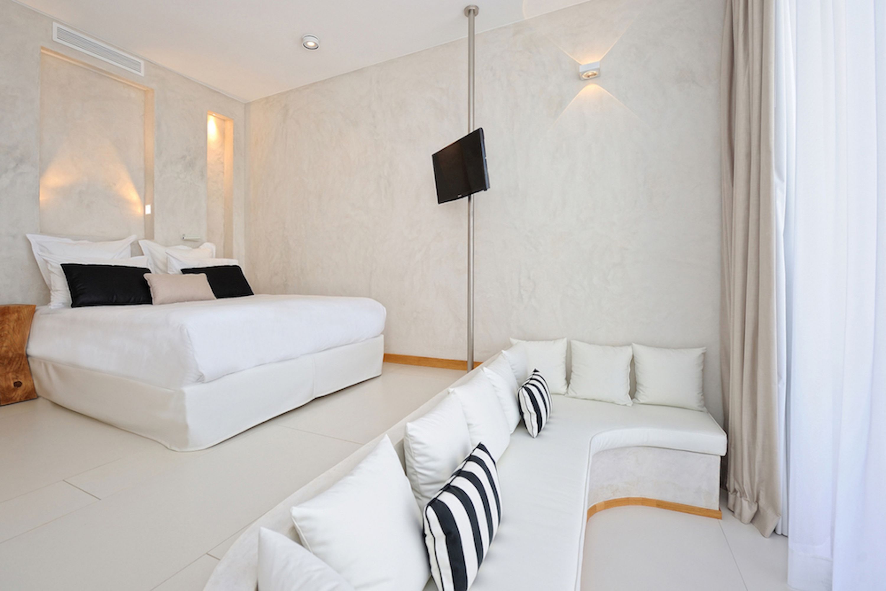 Una habitación de hotel moderna con una cama grande, un sofá, un televisor de pantalla plana en la pared y un amplio ventanal con cortinas.