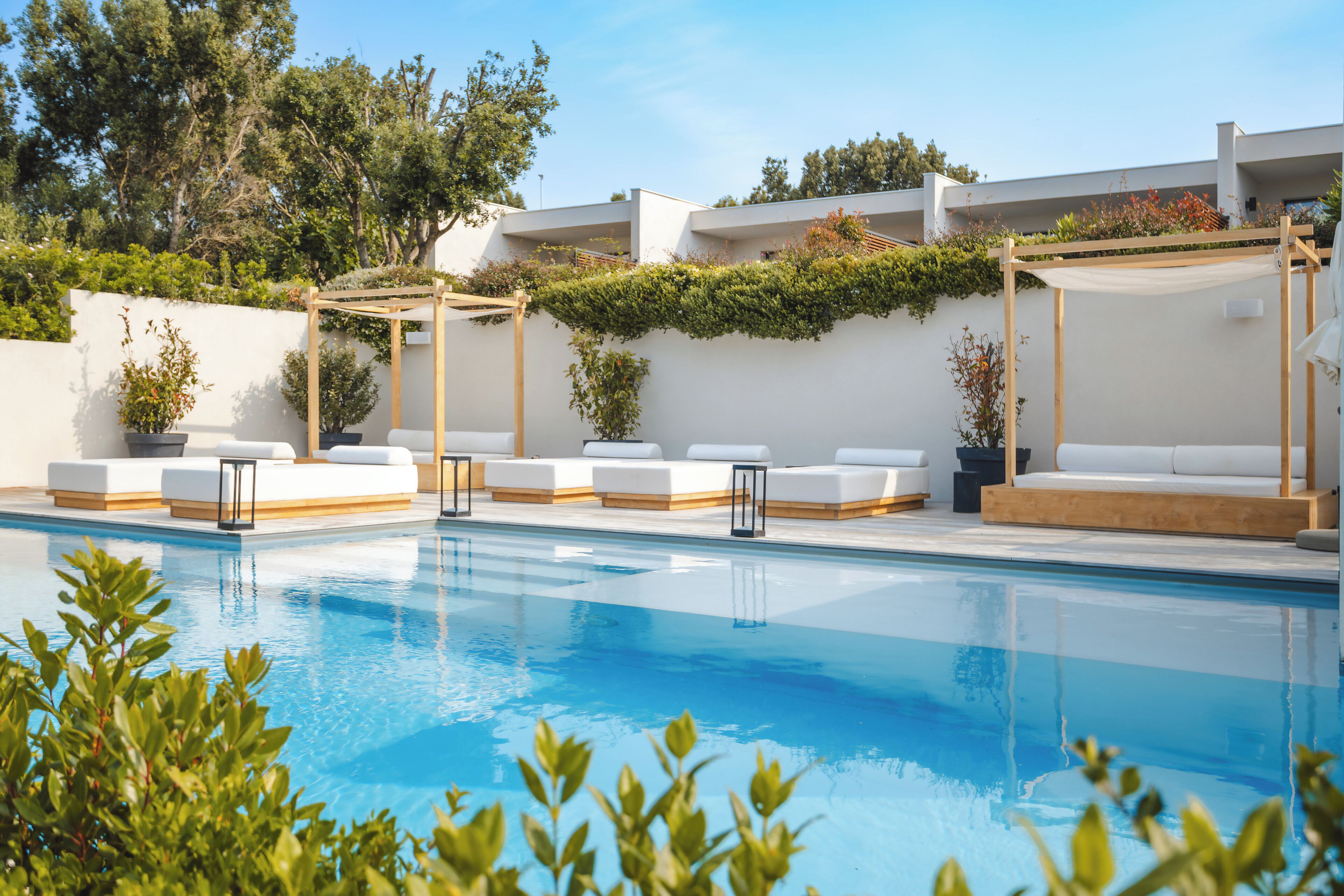 Piscina con sillas de descanso blancas y una pérgola, rodeada de follaje y un edificio blanco.