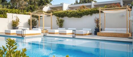 Piscina con sillas de descanso blancas y una pérgola, rodeada de follaje y un edificio blanco.