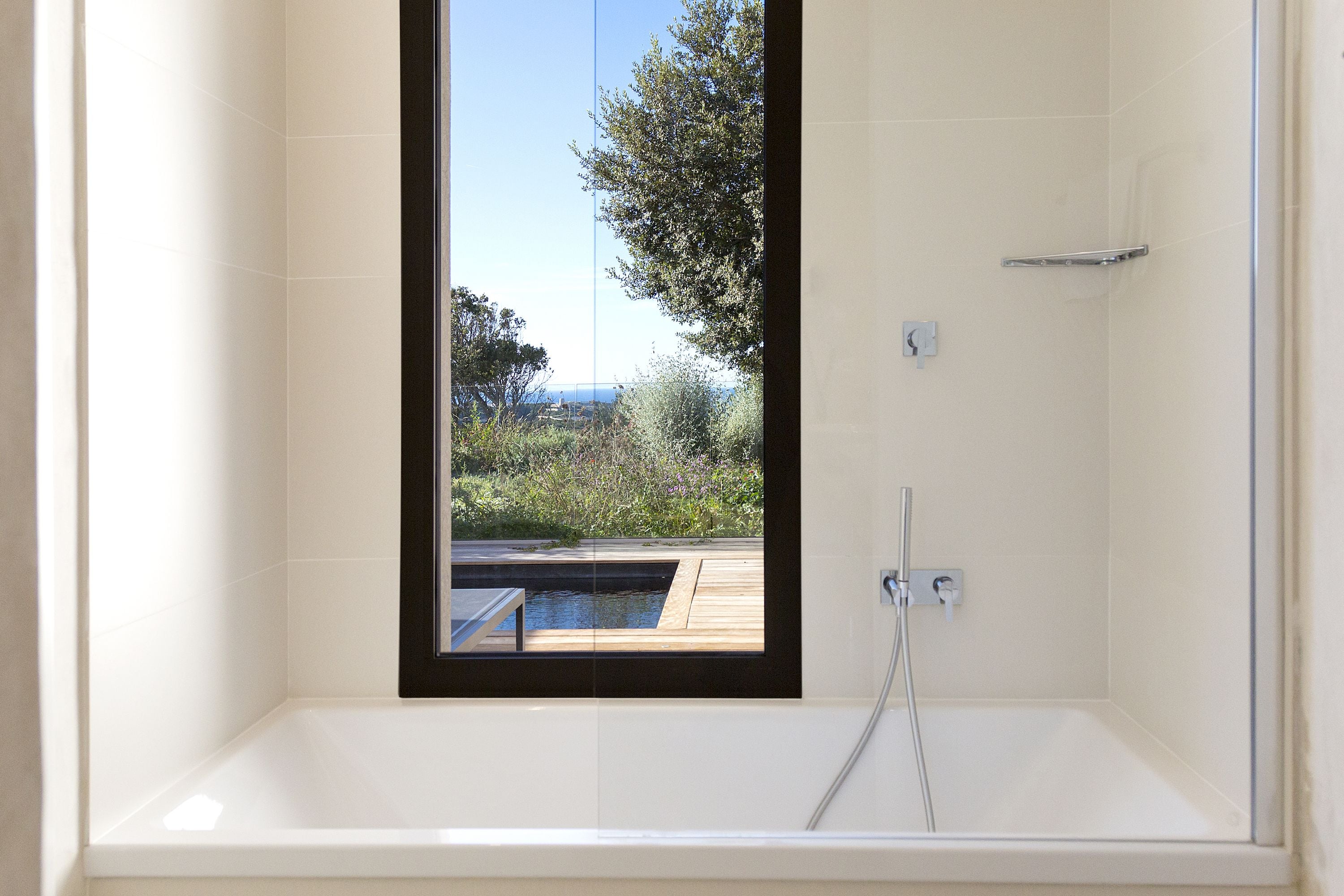 A modern bathroom with a large bathtub and a window overlooking a pool and greenery.