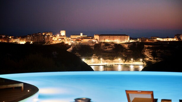An infinity pool with a view of a historic cityscape at night. - Hotel Cala di Greco (Bonifacio)