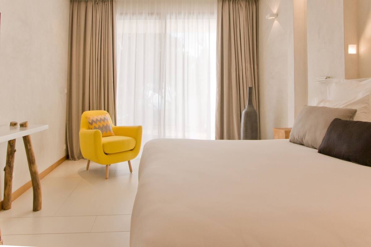 A hotel room with a large bed, a yellow armchair, a small table, and a window with curtains.