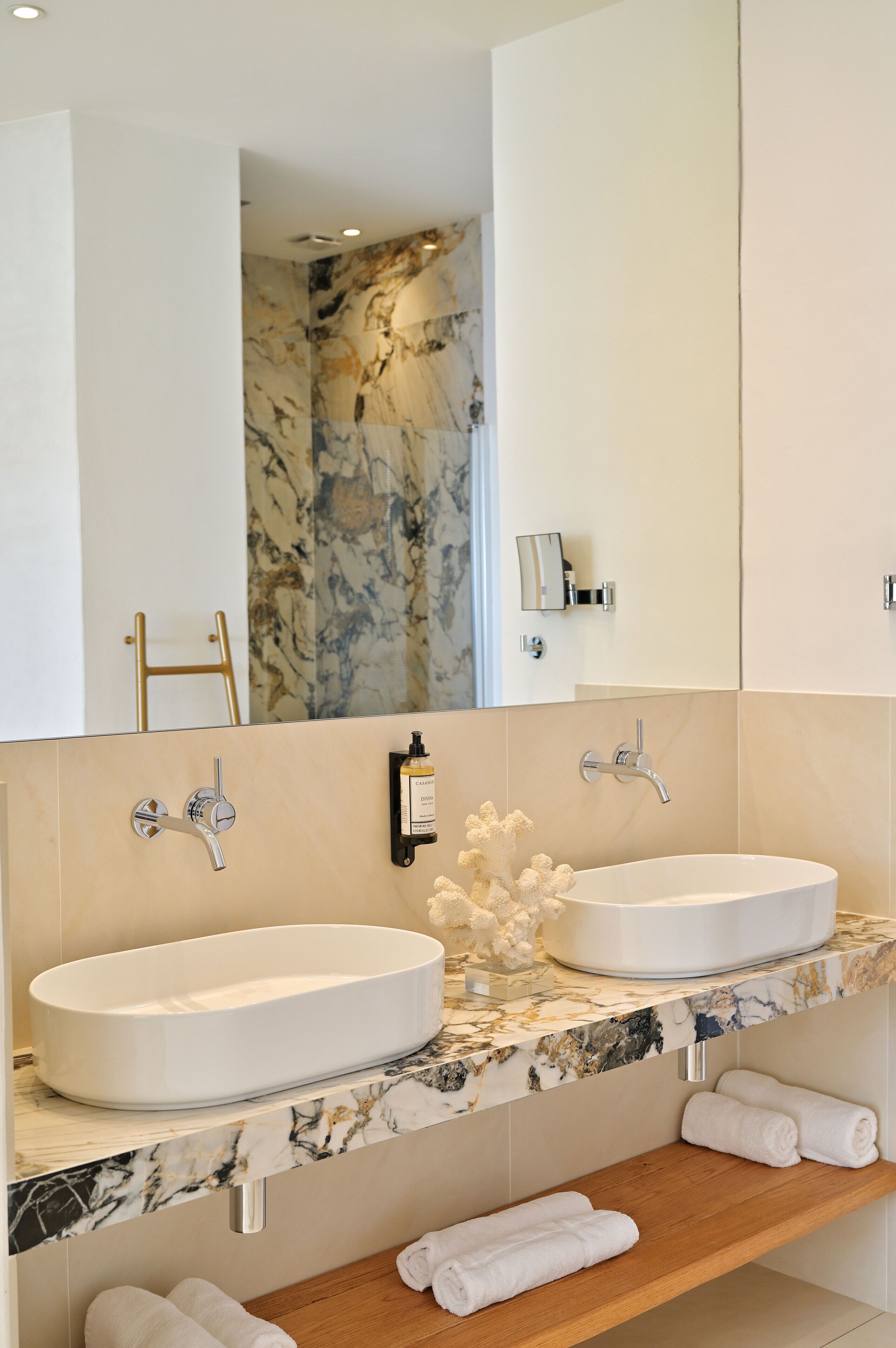 A modern bathroom with two sinks, marble countertops, and a wooden shelf with towels.
