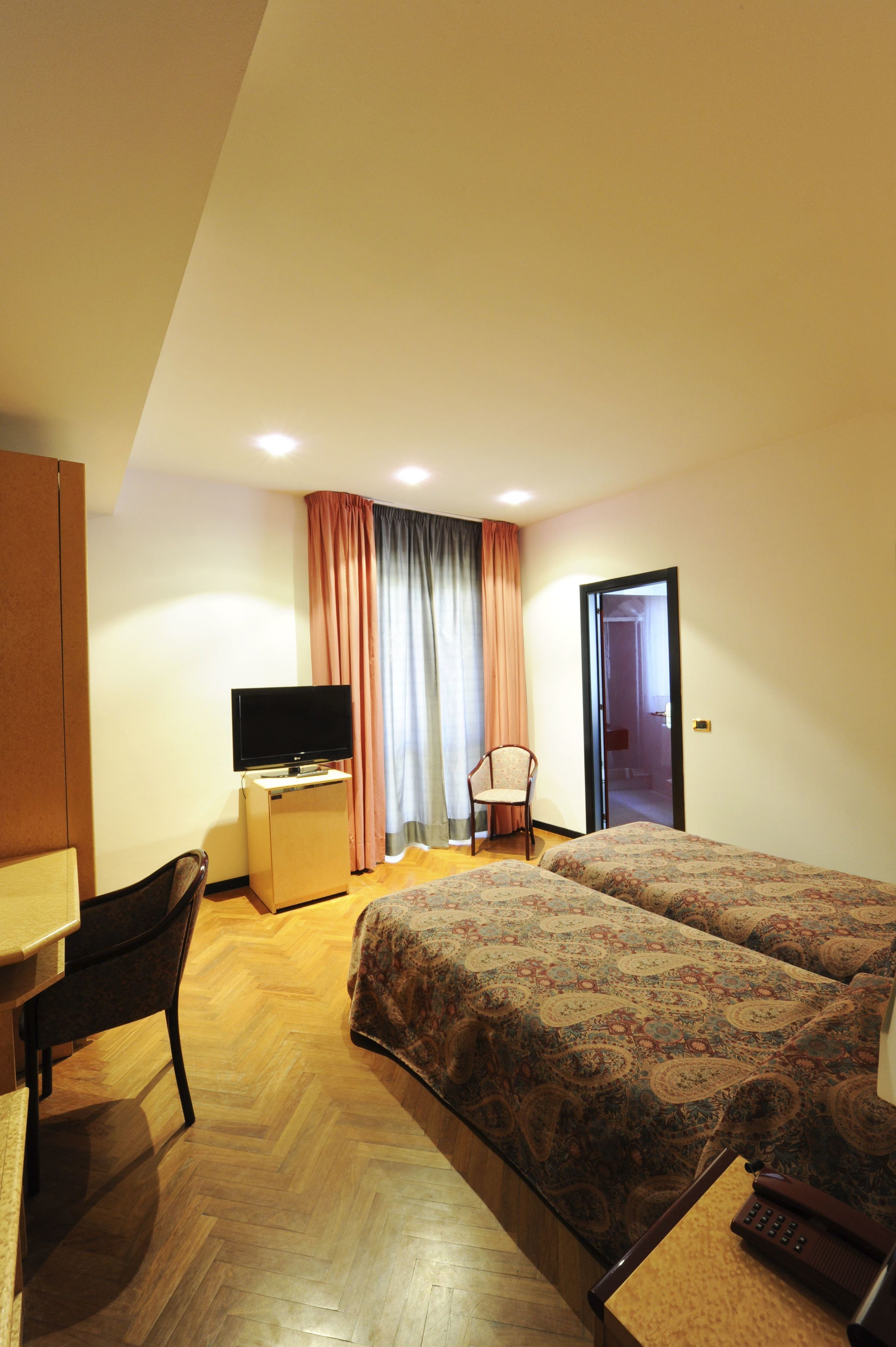 A hotel room with two beds, a desk with a TV, a chair, and a door leading to another room.