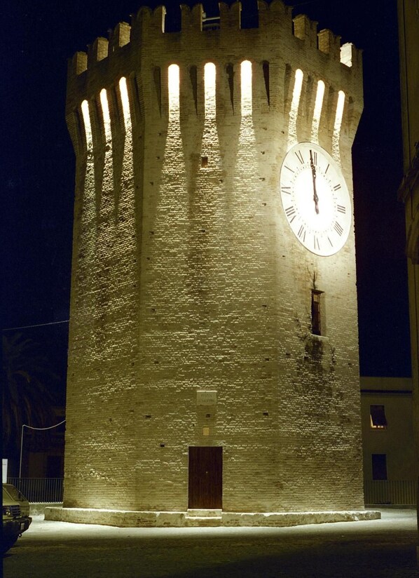 Una torre con un reloj en la parte superior, una puerta pequeña en la base y un área pavimentada al frente.