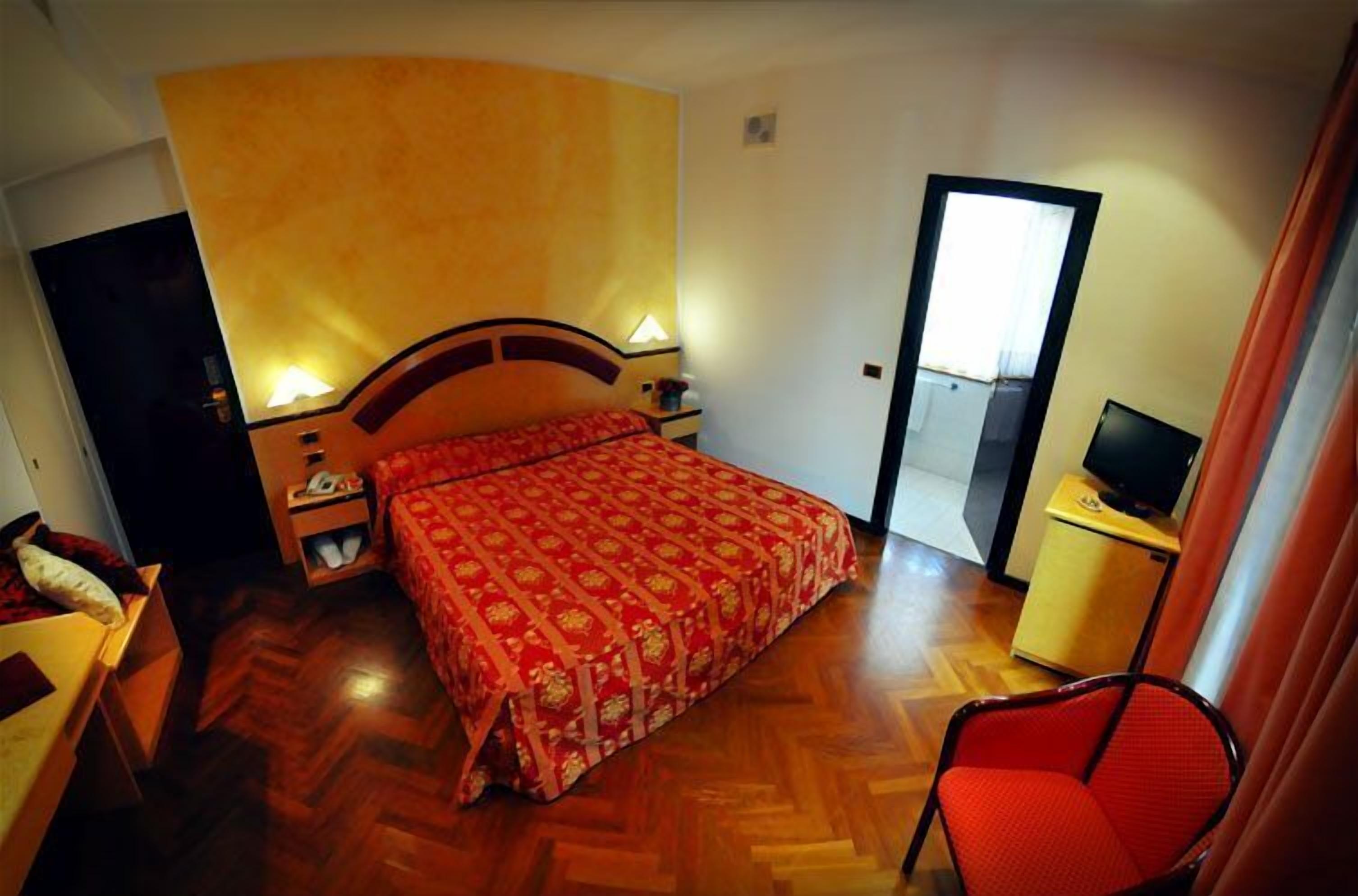 Ein Hotelzimmer mit Holzboden, einem Bett mit rotem, gemustertem Überwurf, einem Nachttisch mit Lampe, einem Fernseher auf einem kleinen Tisch und einem Sessel.