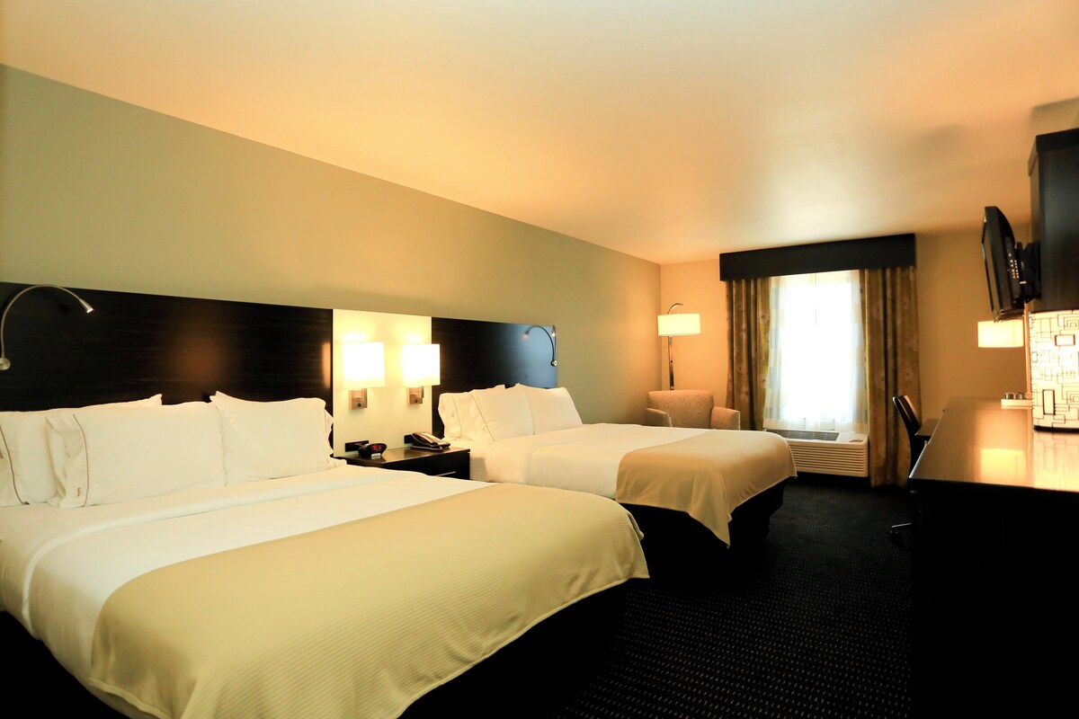 A hotel room with two beds, a desk, a chair, a TV, and a window with curtains.
