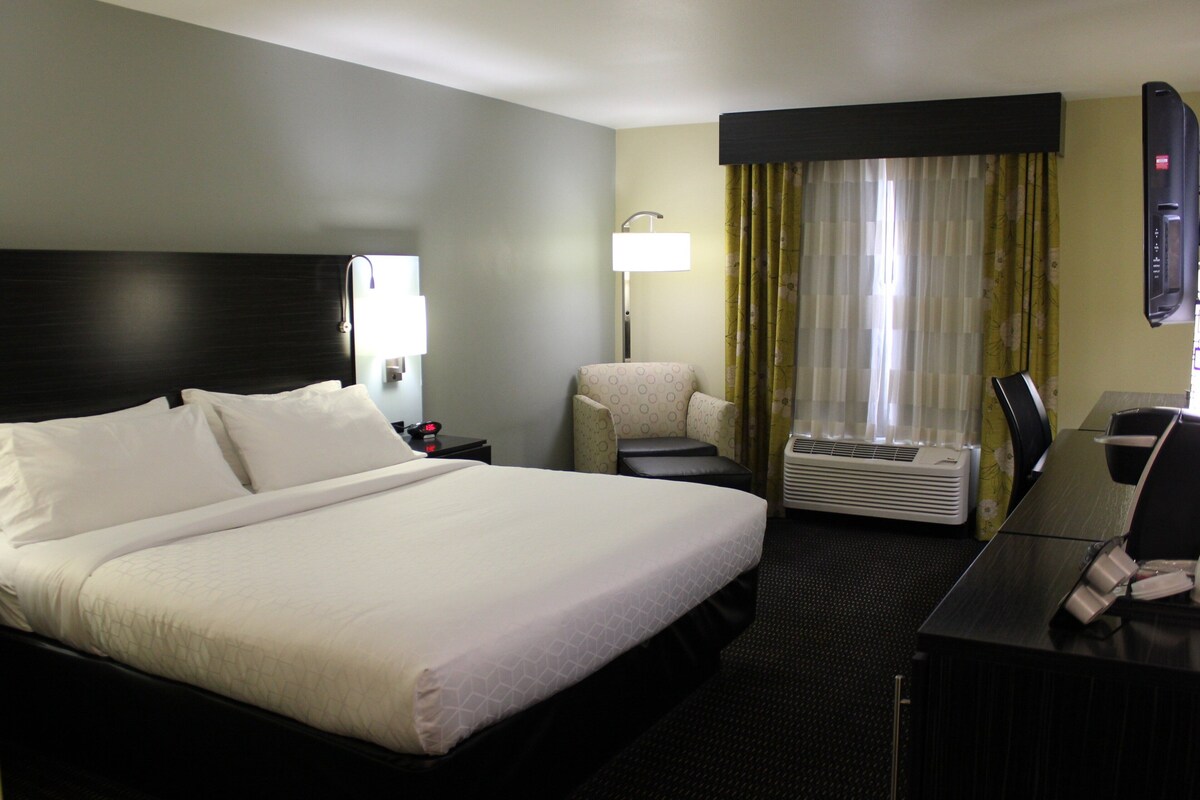 A hotel room with a large bed, a desk with a chair, a lamp, and a window with curtains.