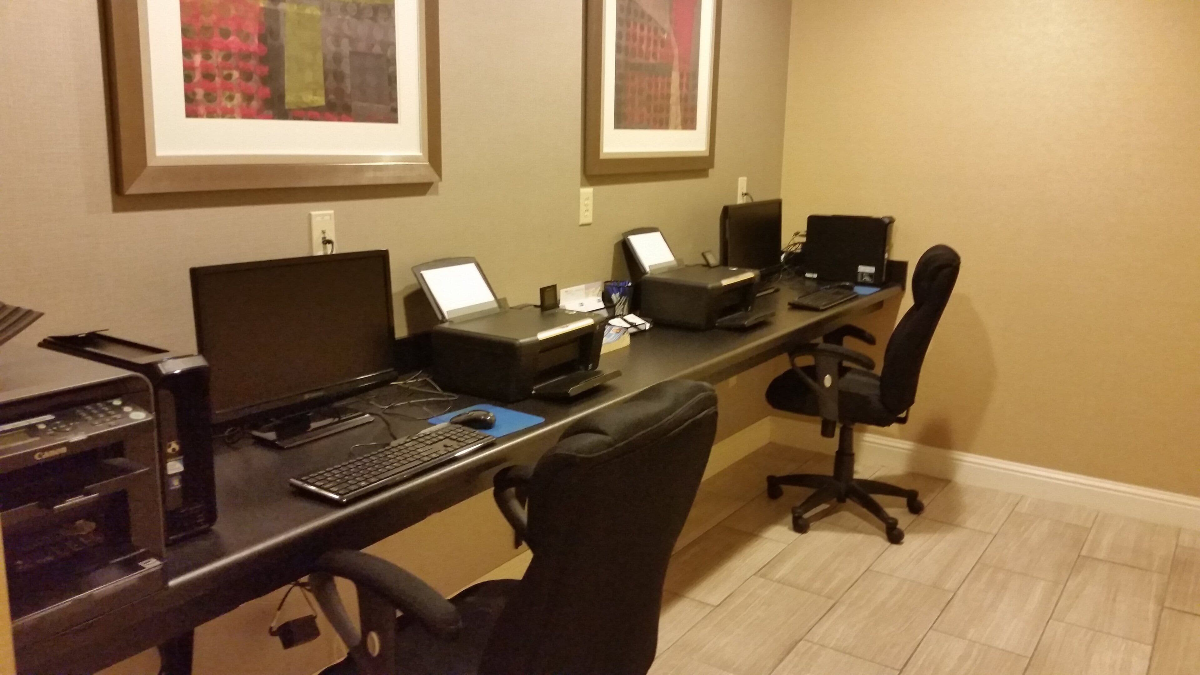 Habitación de hotel con un escritorio equipado con un ordenador de sobremesa, una impresora y un teclado. Hay dos monitores, una silla y un cuadro en la pared.
