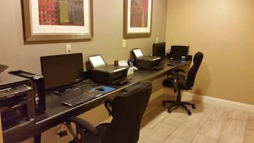 Habitación de hotel con un escritorio equipado con un ordenador de sobremesa, una impresora y un teclado. Hay dos monitores, una silla y un cuadro en la pared.