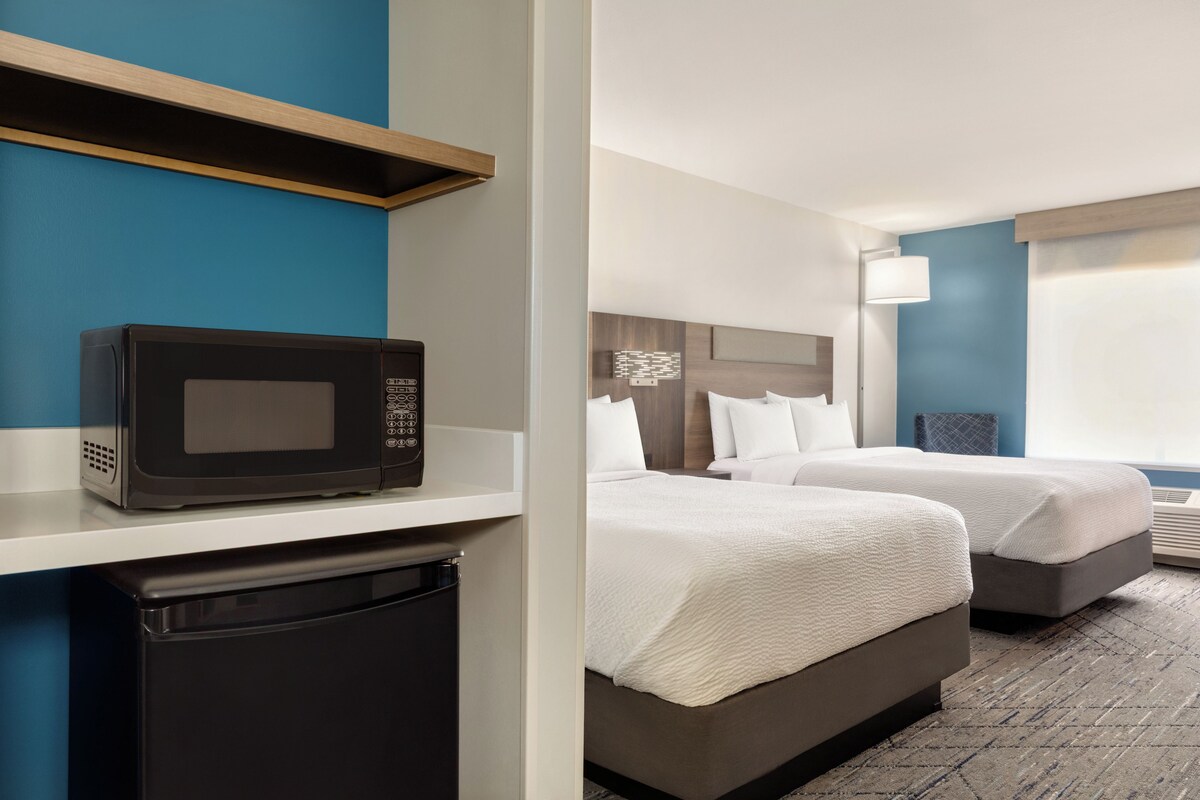 A hotel room with a microwave, a small refrigerator, two beds with white bedding, a nightstand, a lamp, and a window with blinds.
