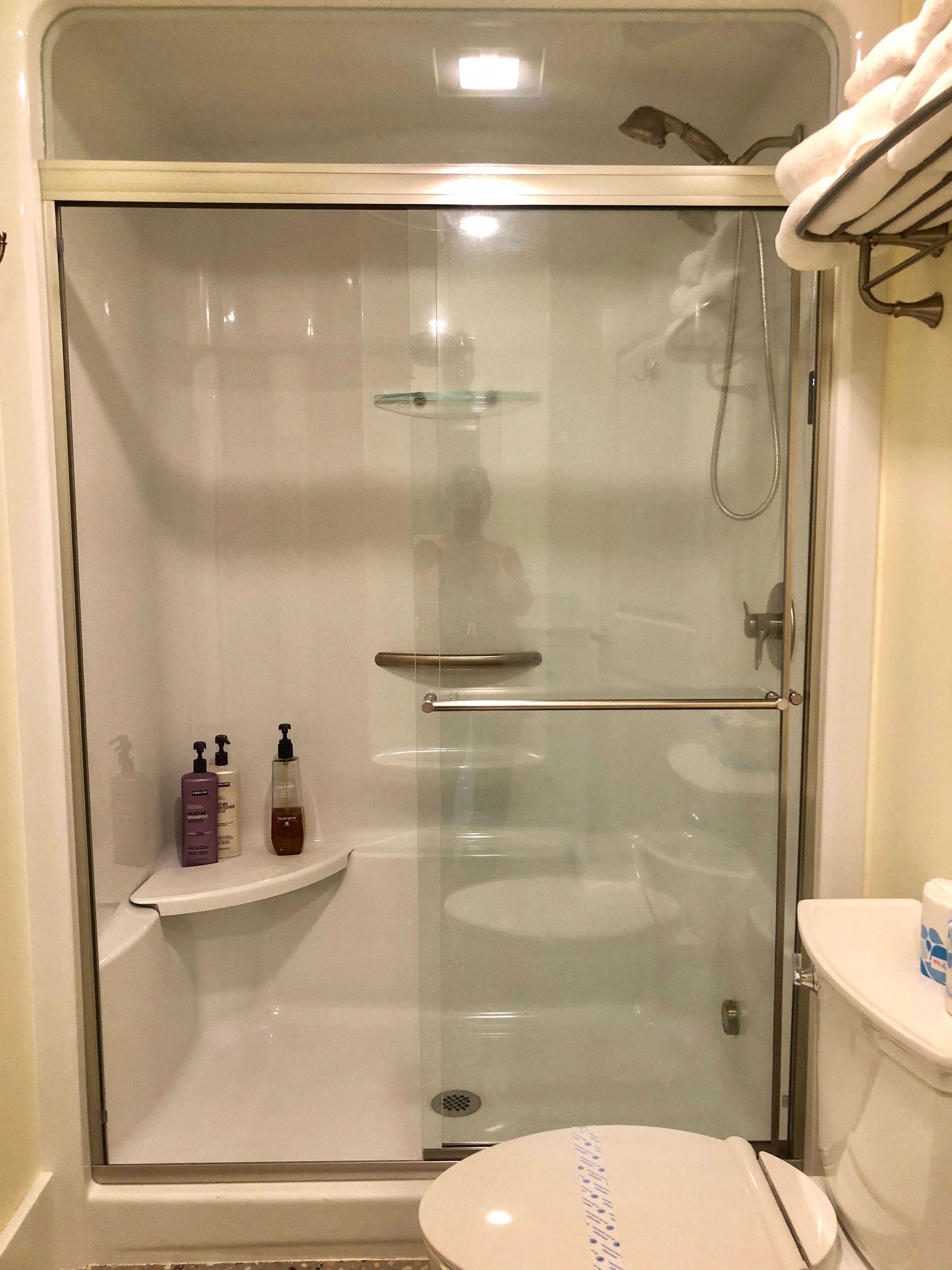 A glass-enclosed shower stall with a glass door, a white toilet, and a shelf with toiletries.