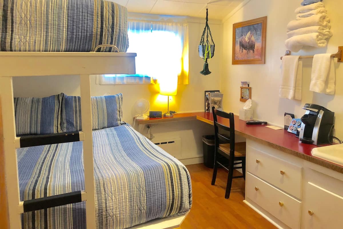 Une chambre d’hôtel compacte comprenant un lit superposé, un bureau avec une chaise, une petite kitchenette et une fenêtre avec des rideaux.