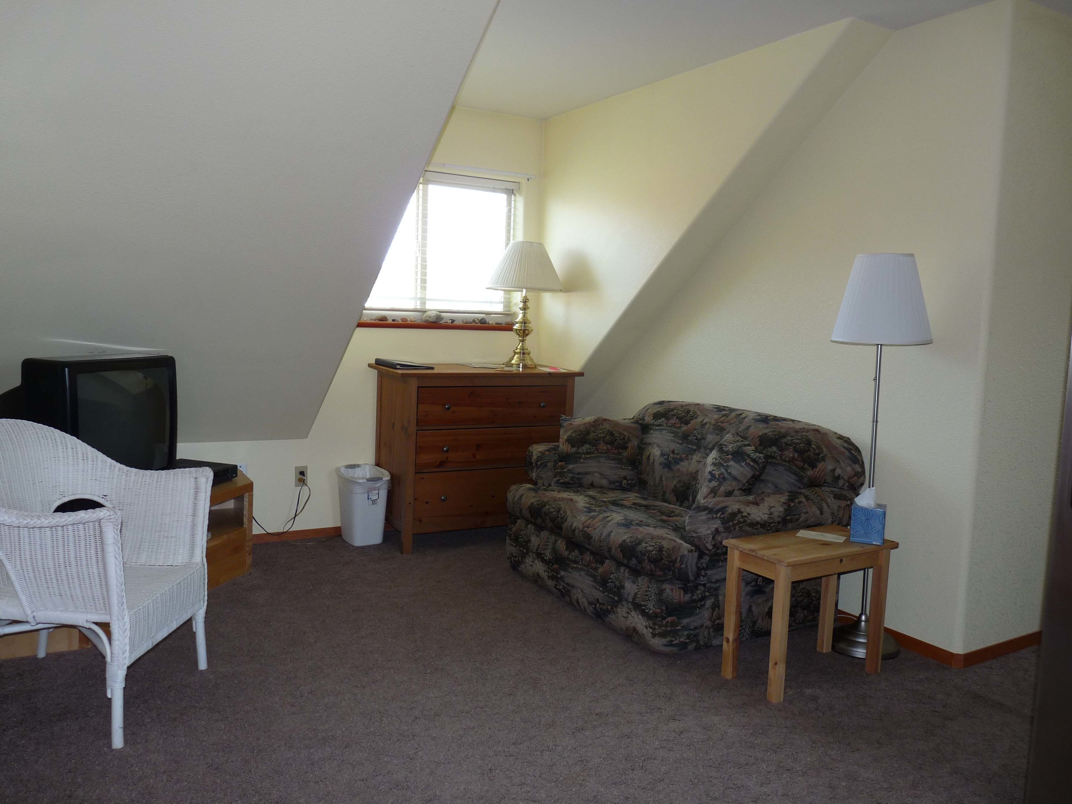 Una habitación pequeña y con techo inclinado, con un televisor, un sillón de mimbre blanco, un sofá estampado, un aparador de madera y una lámpara.