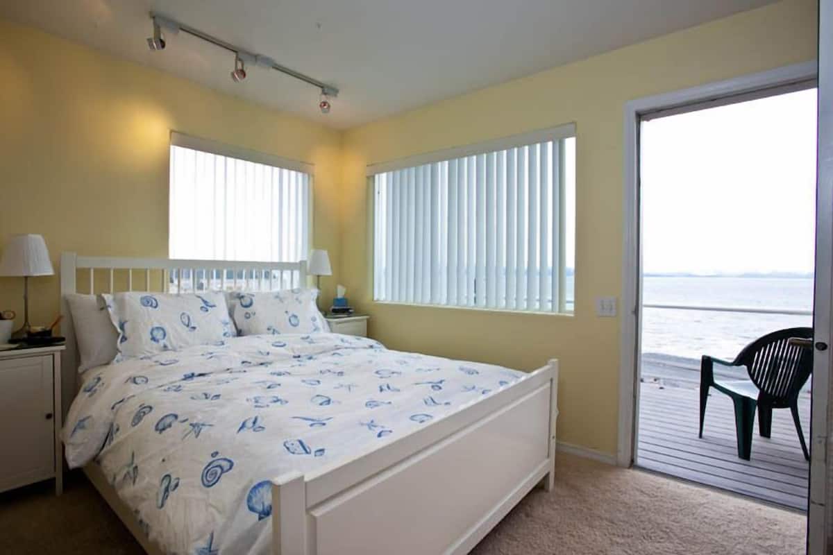 Une chambre à coucher avec un lit, deux tables de chevet, une fenêtre avec des stores et une vue sur l’océan, visible à travers une porte.