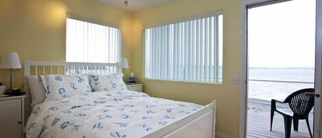 A bedroom with a bed, two bedside tables, a window with blinds, and a view of the ocean through a door.