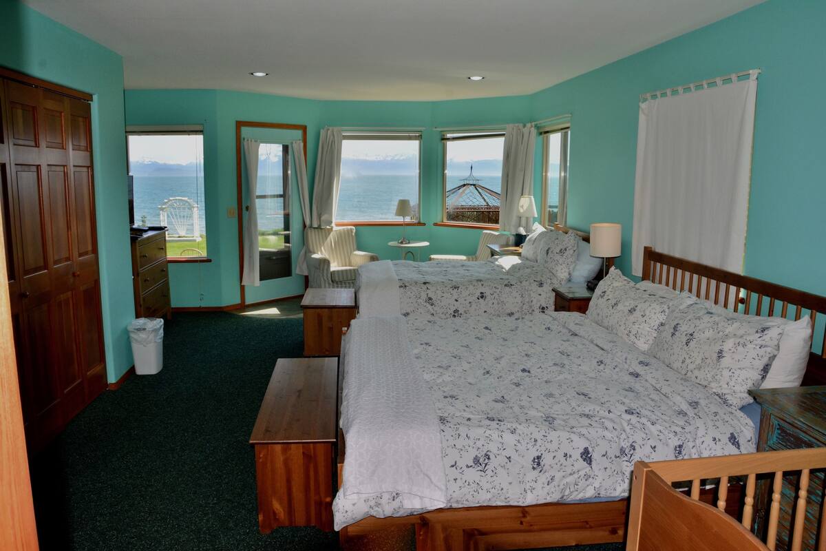 Une chambre avec un grand lit, une tête de lit en bois, une table de chevet, un bureau et une chaise. Il y a deux fenêtres avec des rideaux, une vue sur la mer et les montagnes, et un petit balcon avec une table et des chaises.