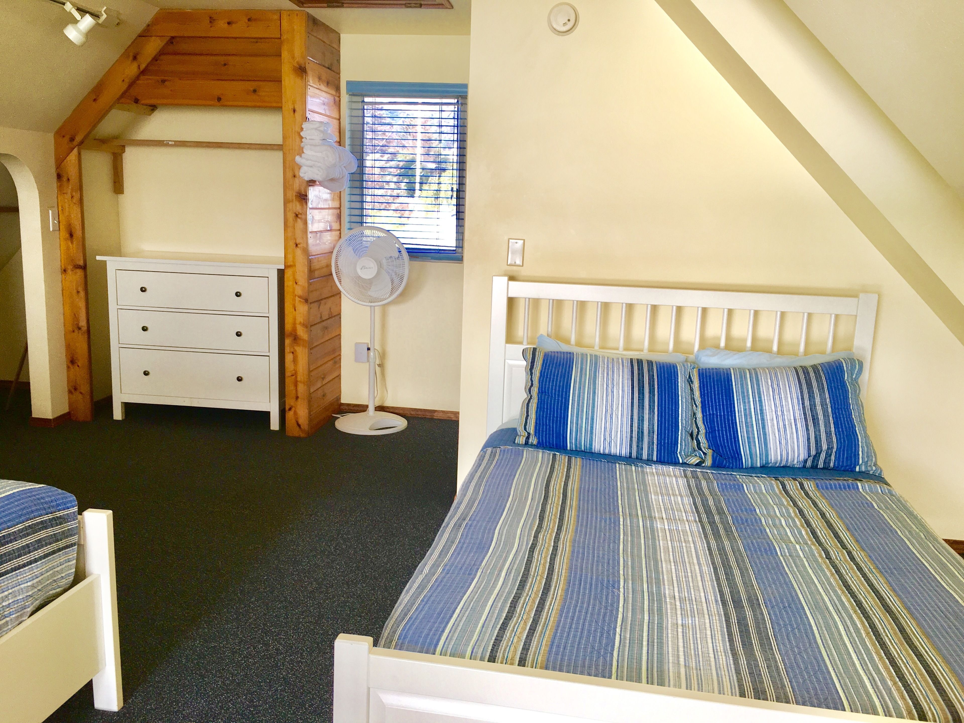 Un dormitorio con techo inclinado, una cama con ropa de cama a rayas, un aparador blanco y un ventilador de pie.