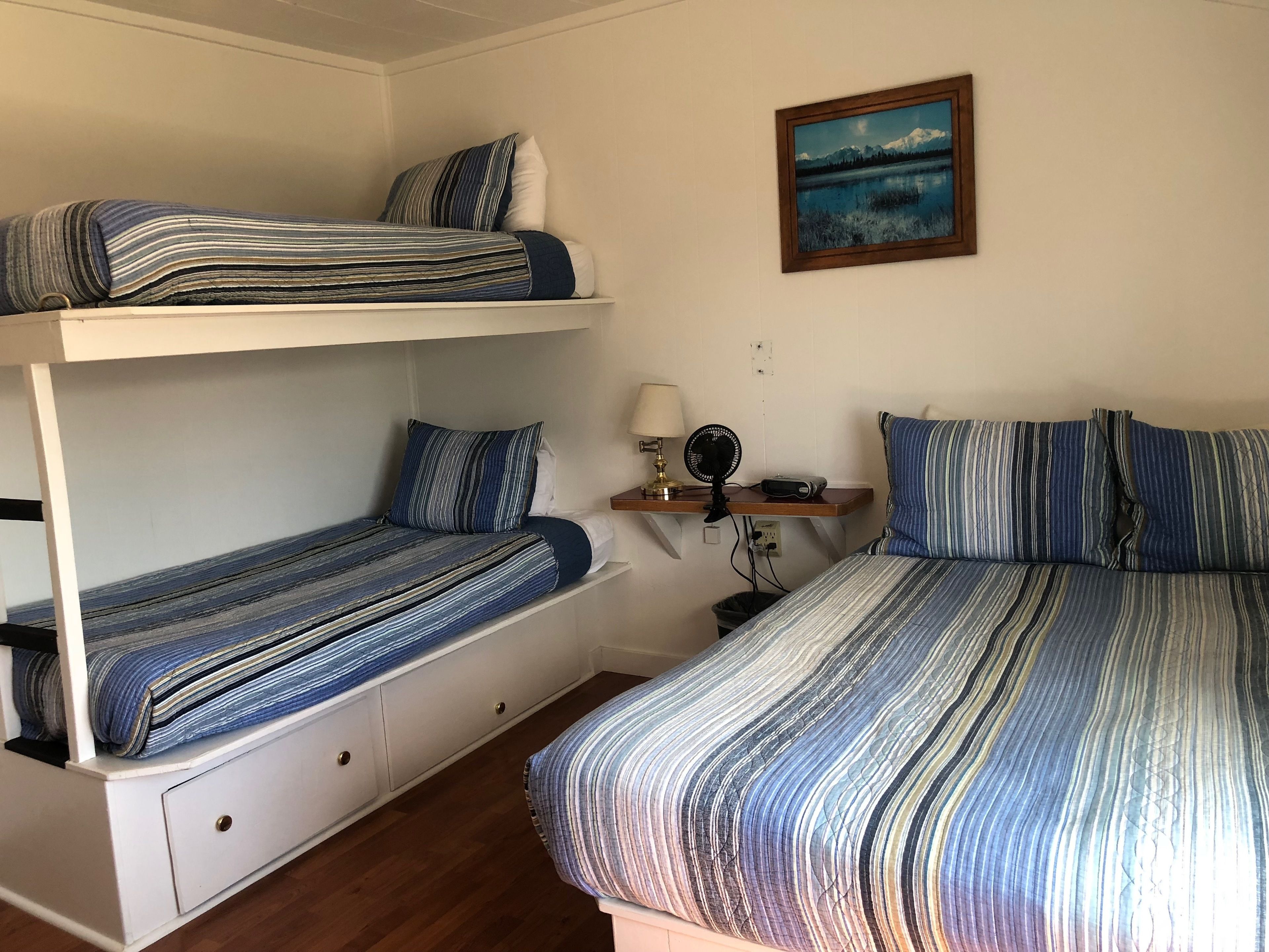 Una habitación con literas, un escritorio con lámpara, un ventilador y un cuadro en la pared.