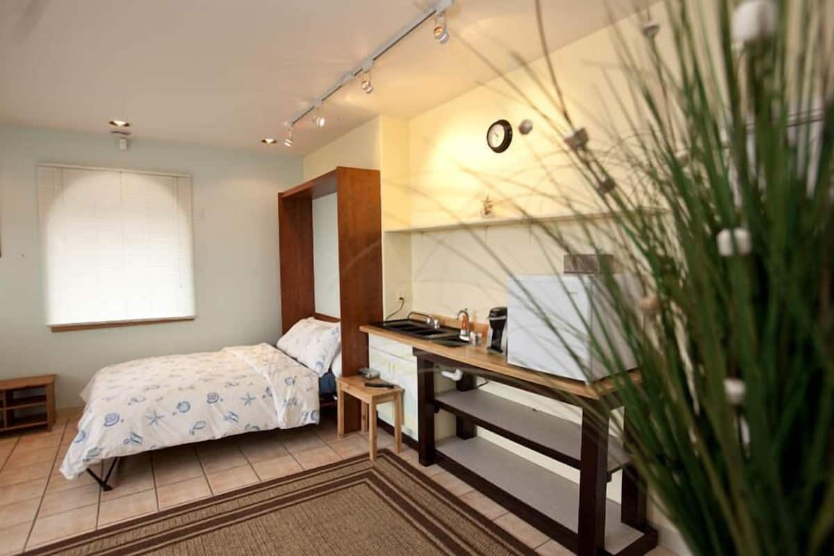 Une chambre à coucher comprenant un lit, un bureau, une chaise, une petite table, un radiateur, une horloge et une fenêtre avec des stores.