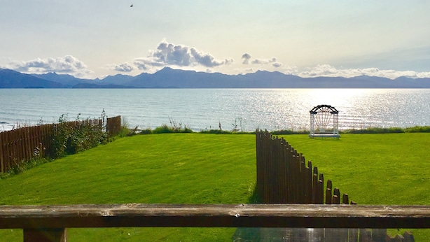 A grassy area with a wooden fence, a gazebo, and a body of water with mountains in the background. - Driftwood Inn and Seaside Lodges (Homer)