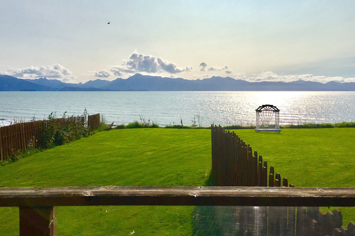 Une zone herbeuse, délimitée par une clôture en bois, avec un belvédère situé au bord d’un lac, des montagnes se dressant à l’horizon.