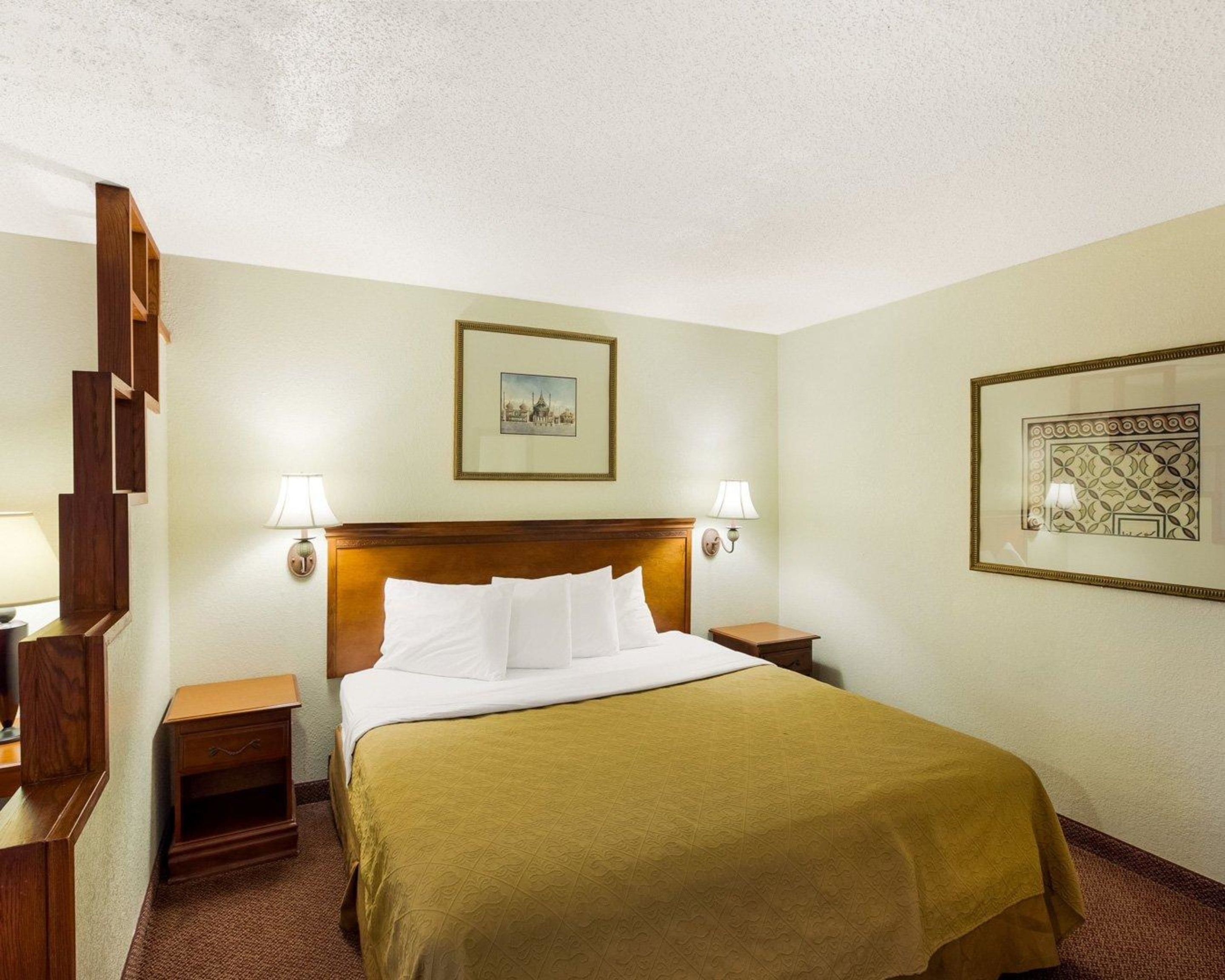 Habitación de hotel con cama, dos mesitas de noche, cabecera, dos lámparas de pared y dos cuadros enmarcados.