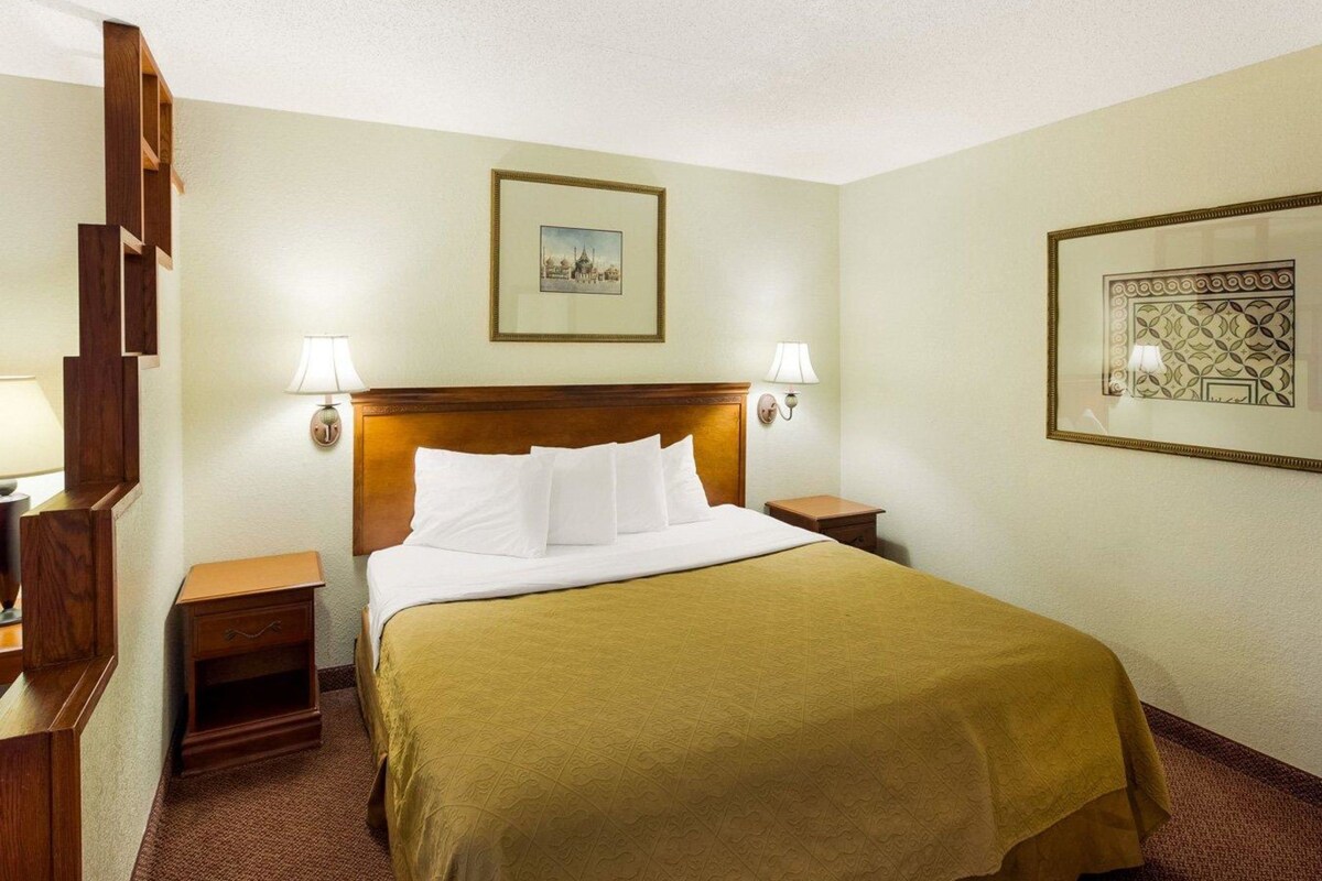 A hotel room with a bed, two bedside tables, a headboard, two wall-mounted lamps, and two framed pictures.