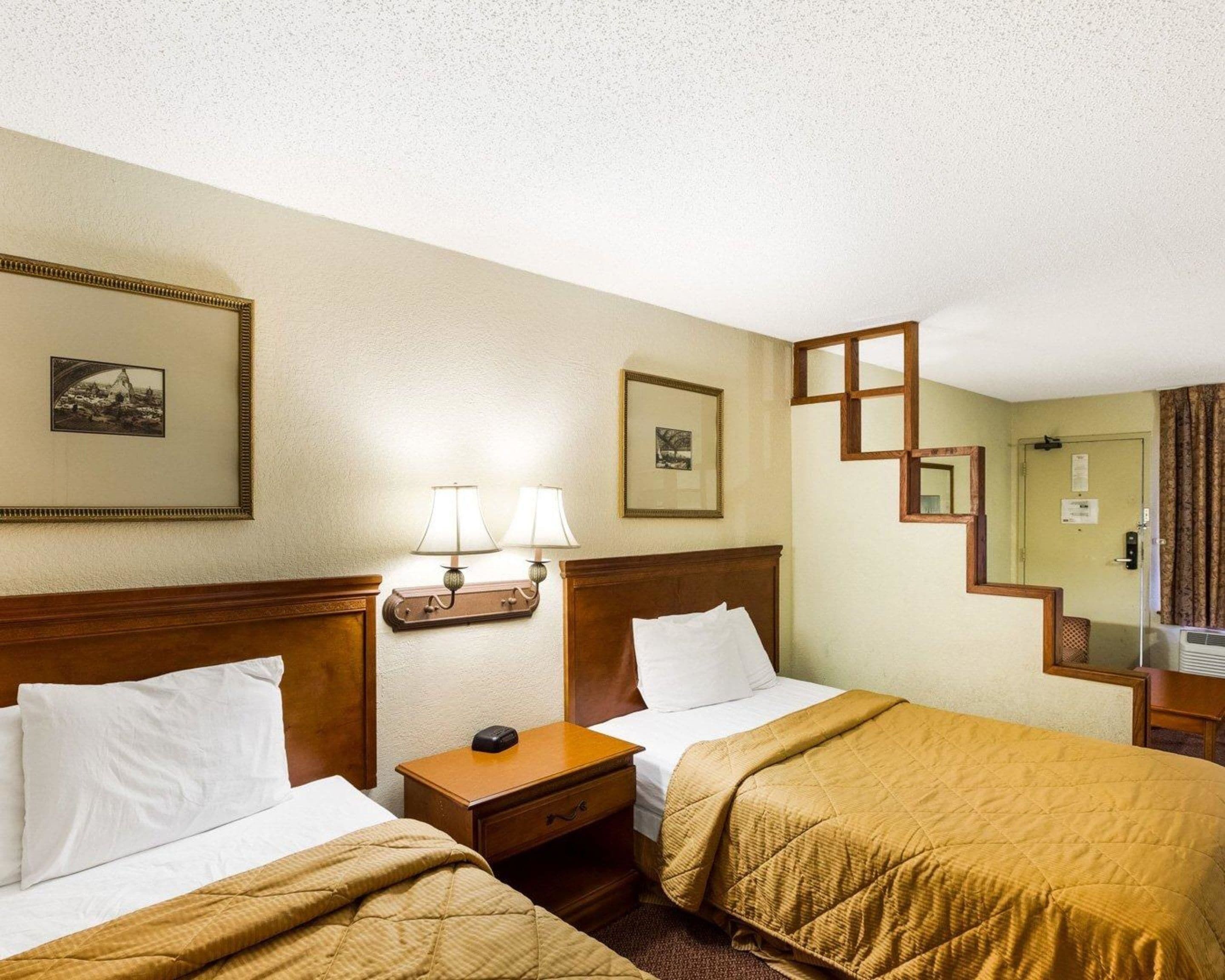 Una habitación de hotel con dos camas, una mesita de noche, una lámpara, un cuadro enmarco y un espejo.