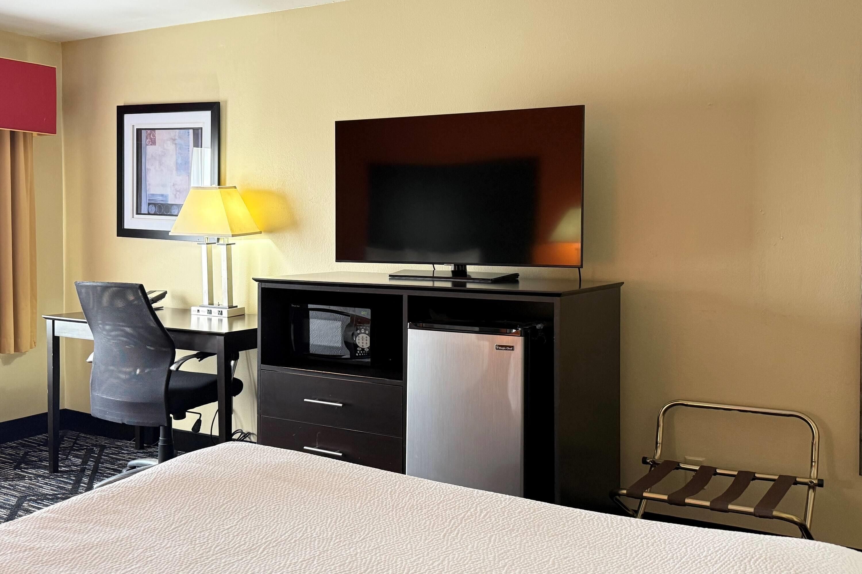 A hotel room with a bed, a desk with a lamp, a television, a refrigerator, and a chair.