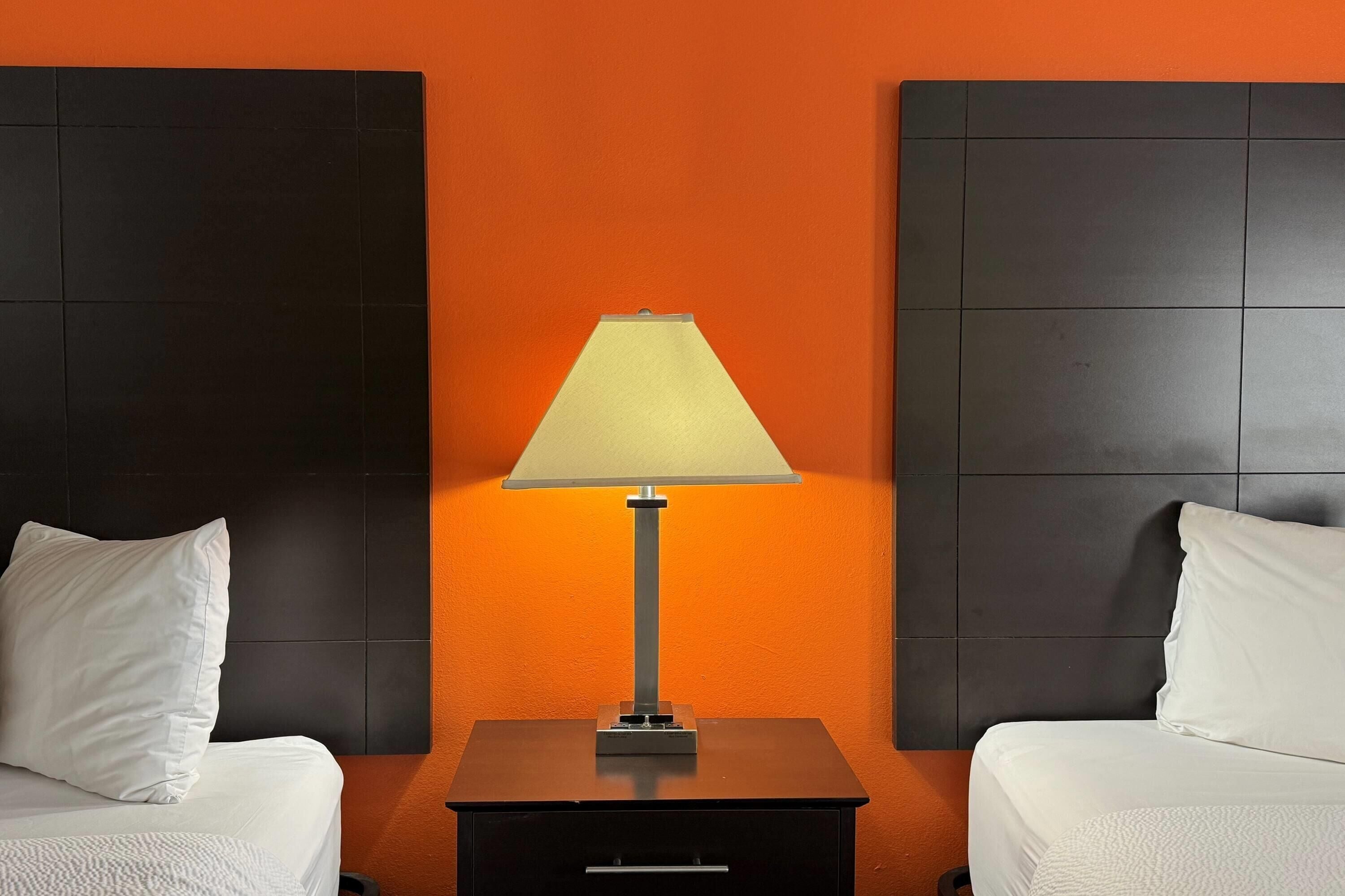 Una camera d'albergo con due letti, un comodino con una lampada e una parete arancione.