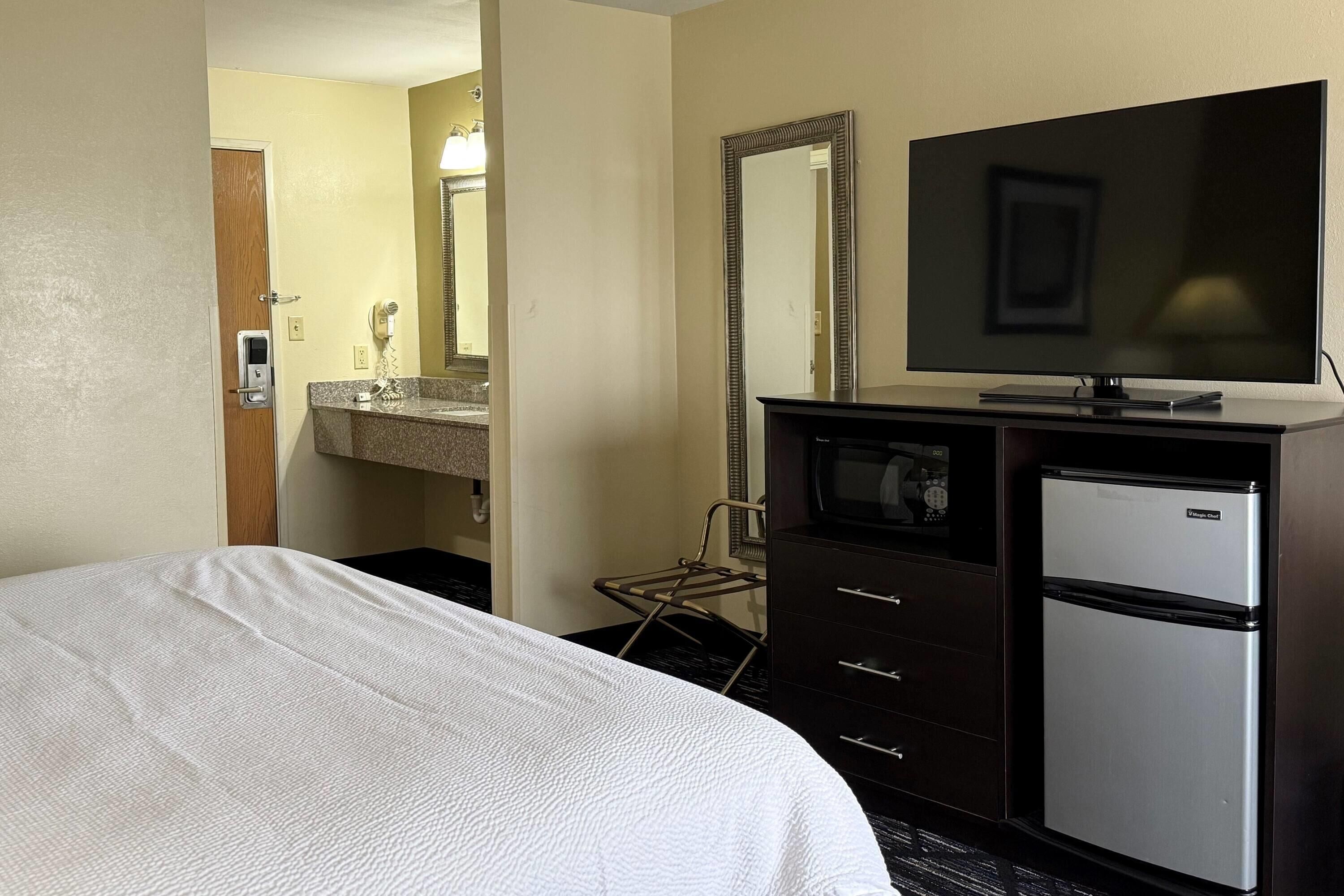 A hotel room with a bed, a dresser, a refrigerator, a television, and a bathroom with a sink and mirror.