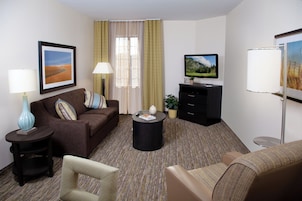 Habitación de hotel con un sofá marrón, una pequeña mesa redonda, un televisor, una lámpara y un cuadro en la pared.