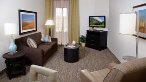 Een hotelkamer met een bruine bank, een klein rond tafeltje, een televisie, een lamp en een raam met gordijnen.