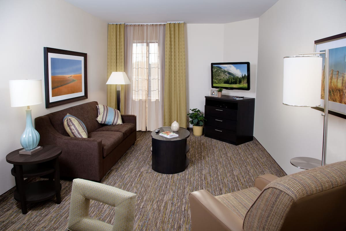A hotel room with a brown sofa, a small round table, a television, a lamp, and a window with curtains.