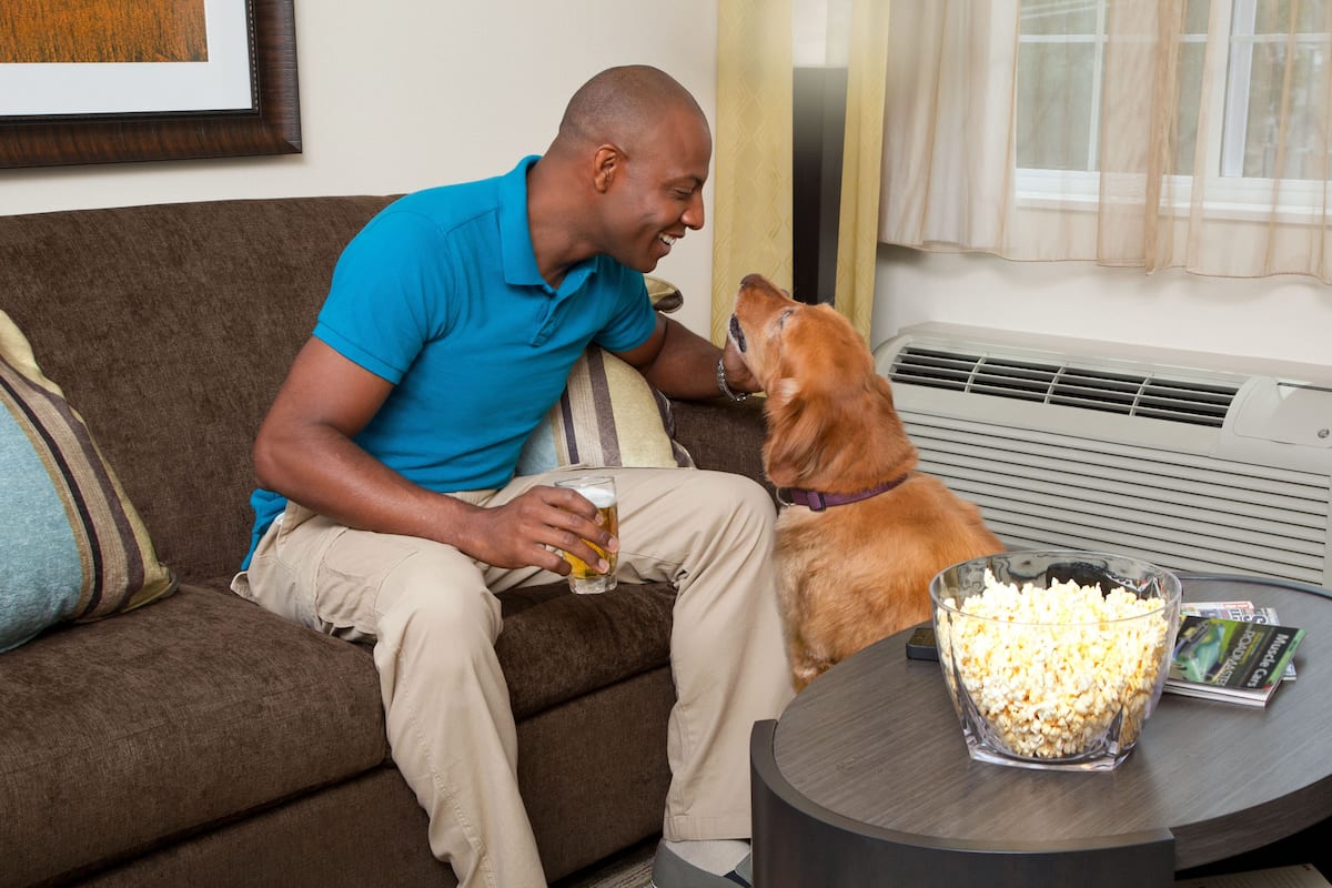 A man sitting on a couch with a dog, holding a glass of beer, and a bowl of popcorn on a table.