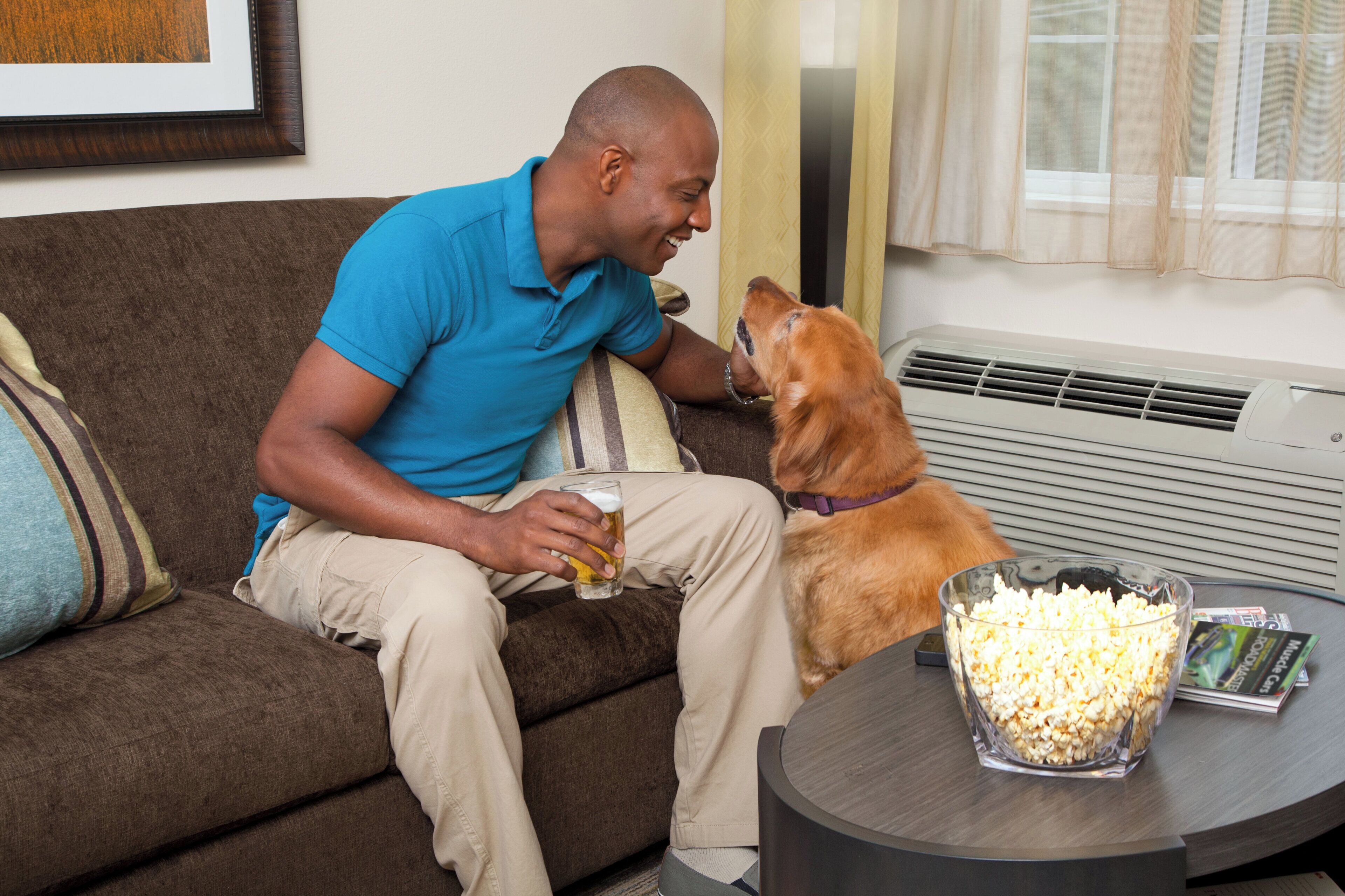 A man sitting on a couch with a dog, holding a glass of beer, and popcorn on a table.